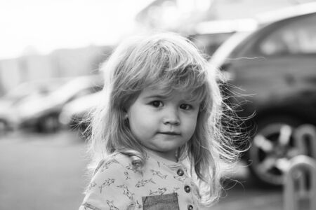 Cute baby boy with long blond hair walking on city street on sunny summer day outdoors on blurred...