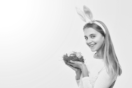 happy easter girl in pink bunny ears with colorful painted eggs in straw nest, has long blonde hair and smiling adorable face isolated on white background. traditional spring holiday food, copy spaceの写真素材