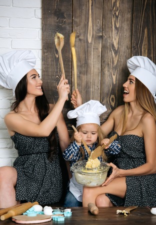 Baby boy kneading dough in bowl. Two women having fun with wooden spoons. Brother and sisters in chef hats in kitchen. Homemade baking and cooking. Happy family and childhood concept.の写真素材