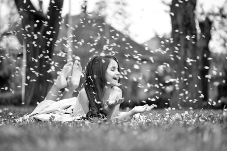 Beautiful little girl in pink dress with long brunette hair and smiling face lying on green grass in spring flower blossom petals outdoorの写真素材