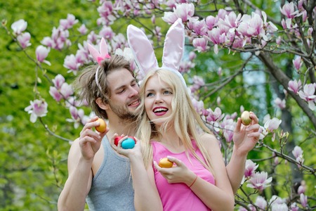girl and handsome man or macho with bunny ears holding colorful eggs on floral environment. Happy girlfriend and boyfriend in blossoming garden. Couple in love. Spring. Easterの写真素材