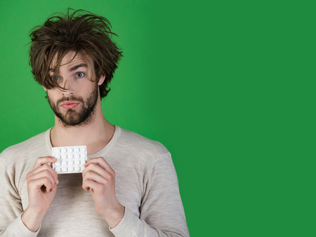 man with pills in blister pack has uncombed hair in morning on green background, health and medicine, handover and headache, drug, cold and flu, insomnia, copy spaceの写真素材