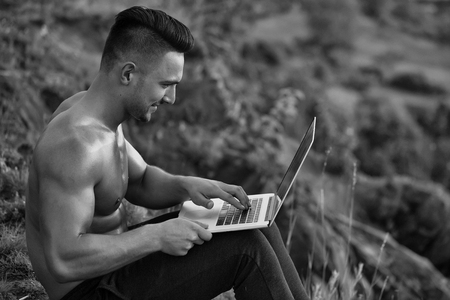 Young handsome smiling man with muscular sexy body and bare chest sitting with laptop outdoor sunny dayの写真素材