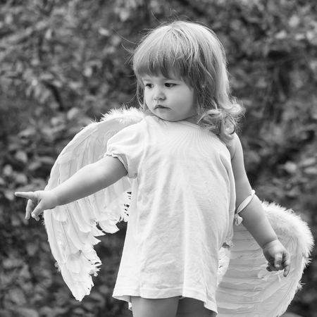 small cute baby boy with blonde long hair in white feathered angel wings and cloth outdoor on green natural backgroundの写真素材