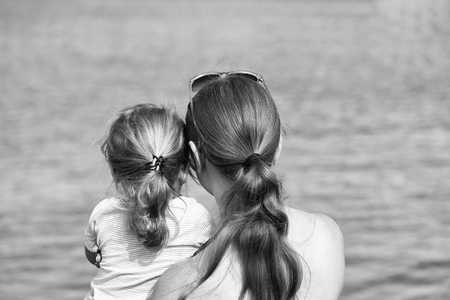 Mother and son, beautiful woman and cute baby boy, with blond hair ponytails look at sea or ocean water on sunny summer day on blurred blue backgroundの写真素材