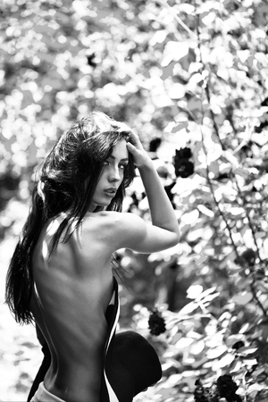 young glamour sexy woman with bright makeup on pretty face and long brunette hair has naked slim body holds red round retro hat near rose flower bush outdoor, closeupの写真素材
