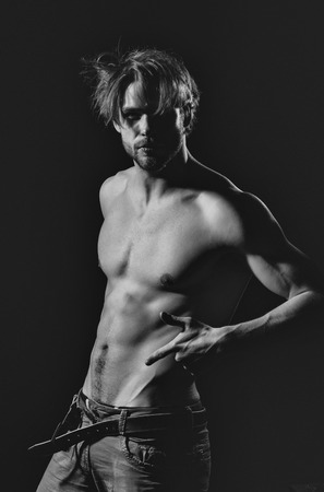 Handsome man or unshaven macho, bodybuilder, with stylish blond hair, haircut, sexy, naked, muscular torso with six packs and abs, biceps, triceps unfastening belt on black backgroundの写真素材