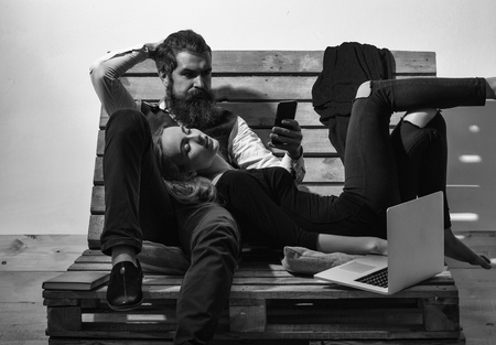 Young couple spend time together with laptop, computer. Pretty girl or beautiful woman sleeps on knees of handsome man, bearded hipster with beard, talking on mobile phone on wooden pallet sofaの写真素材