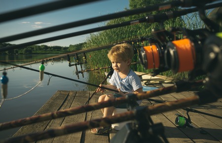 Little baby boy fishing on bank of river with fishing rod in hands.