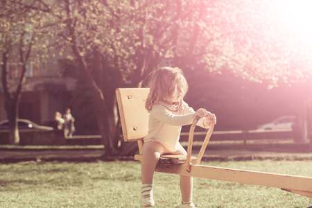 Girl sit on seesaw on sunny day. Child have fun on playground. Kid on teeter totter outdoor. Balance, equilibrium, harmony. Childhood, activity, lifestyle.の写真素材