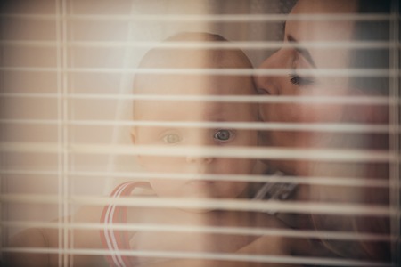 Mother kiss baby son at window shutters. Woman with child with blinds on foreground. Mothers day concept. Family, love, trust.の写真素材