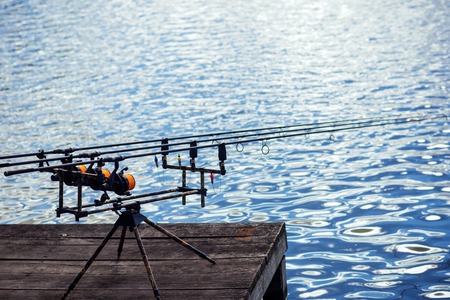 Spinning tackles on pod on wooden pier. Rods and reels at river or lake water. Fishing, adventure, sport, activity. Spin fishing, angling, catching fish. Summer vacation, pastime, hobby.の写真素材