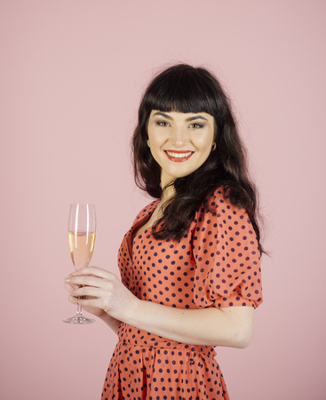 Woman with makeup celebrates with glass of champagne. Brunette girl in dress with polka dots on pink background. Woman with smiling face posing in pinup style. Holidays and celebration concept.の写真素材