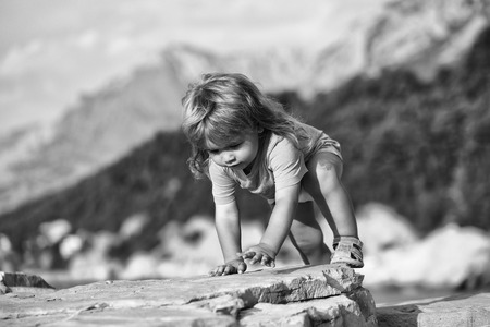 Cute baby boy child with curly blond hair in green shirt plays at rocky sea beach on summer dayの写真素材