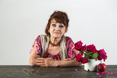 Old lady pose with flowers, apple, glasses on table. Woman with wrinkled face, makeup, beauty, skin aging. Anti aging, rejuvenation, lifting. Beauty, skin aging, skincare. Womens day, 8 march, springの写真素材