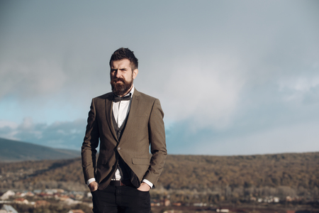 Man with beard and mustache with scenery on background. Hipster with stylish appearance in front of dramatic sky, skyline. Guy with strict face in classic suit with bow tie. Stylish hipster concept.の写真素材