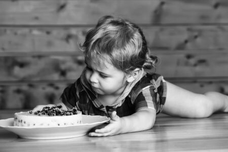 Cute baby boy child with blond curly hair eats delicious cake with blueberries lying on wooden tableの写真素材