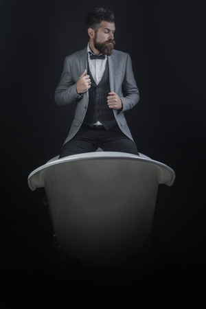 High quality sanitary ware concept. Hipster with stylish appearance in luxury old fashioned bathtub. Man or seller of sanitary ware on dark background. Guy with strict face in suit sits on bathtub.の写真素材
