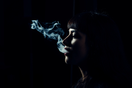 Womans head with smoke in mouth on black background. Side view and shape of female face and smoke. Smoke clouds come out of female lips, copy space. Smoking woman concept.の写真素材