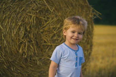 Child play on farm or ranch field, vacation. Little boy smile at hay bale, summer. Summer, harvest season. Agritourism, eco tourism, vacation, travelling. Happy childhood, youth, growth.の写真素材