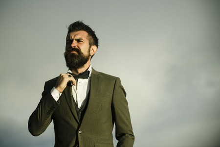 Man with beard and mustache with dramatic sky on background. Stylish hipster concept. Hipster with stylish appearance in front of sky. Guy with thoughtful face in luxury classic suit touches bow tie.の写真素材