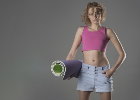 Girl with untidy hair with roll carpet for yoga. Yoga trainer concept. Woman coach with calm face holds yoga carpet ?n grey background, copy space. Sportive lady or fitness trainer in sportive shape.の写真素材