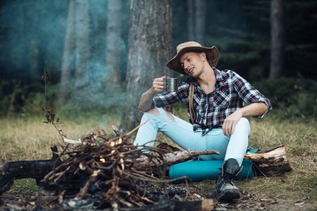 Tourism and hiking concept. Man hiking with overnight stay or picnic. Guy in hat sits near bonfire , trees on background, defocused. Tourist with smiling face relaxing and drinking out of iron mug.の写真素材