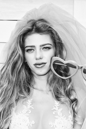 young sexy woman with long blonde hair and smiling pretty face in wedding white dress and bride blue veil holding pink glasses in heart shape on wooden backgroundの写真素材