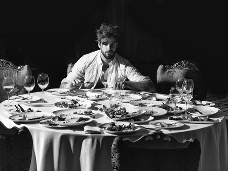 Handsome man with beard and blond messy hair eats with fork and knife at table with leftovers or residues food on dirty plates after banquet dinner in restaurant on dark backgroundの写真素材