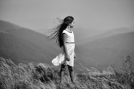 Little cute brunette girl in white lace summer dress looking away standing in mountain valley with deep dry spikelet grass sunny windy day outdoor on natural blue background, horizontal pictureの写真素材