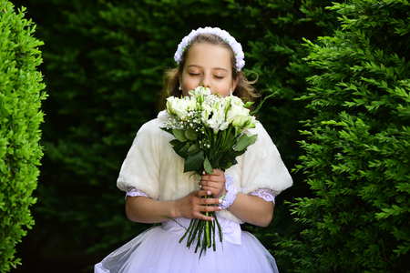 Wedding fashion, beauty salon. Little girl in white dress with rose flower bouquet. Bride girl, bridesmaid and wedding ceremony. Girl child in green summer park, spring. Fashion, Cinderella, princess.の写真素材