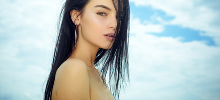 Sensual woman with young face skin, naked shoulders, beauty. Woman with long brunette hair, hairstyle, on cloudy sky. Beauty, look, makeup. Skin care, skincare, treatment, therapy.の写真素材
