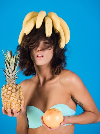 brunette girl in swimsuit with colorful fruit of banana, grapefruit and pineapple on blue background, summer vacation, vitamin and dieting, healthy food and fitnessの写真素材