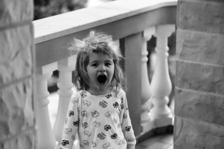 Cute baby boy child with curly blond hair and brown eyes in colorful shirt yawns on balcony after sleepingの写真素材