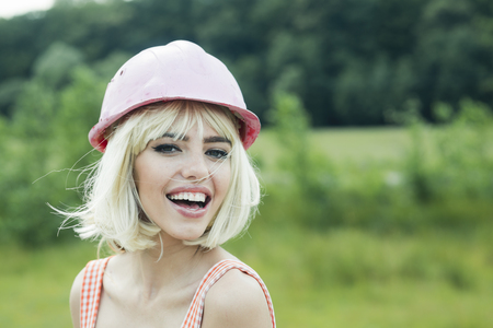 Building and construction, fashion. Sexy woman with blond hair in pink hard hat. Working and repair, glamour girl. Woman with fashionable makeup outdoor, retro. Builder girl in helmet, pinup, beauty.の写真素材