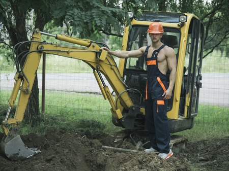 Sexy man with nude torso near construction equipment or excavator on background. Sexy builder concept. Handsome man or bodybuilder with big muscles. Muscular builder in hard hat wears overalls.のeditorial素材