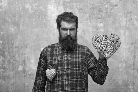Bearded serious man, caucasian hipster, with long beard and moustache in plaid shirt with two hearts, wicker and rosy textile, love gifts for valentines day, on beige grunge wall backgroundの写真素材
