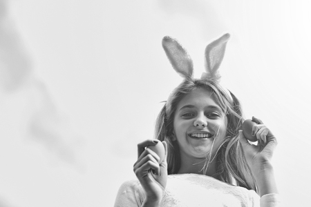 happy girl in pink bunny ears with long blonde hair and smiling adorable face holds colorful painted easter eggs outdoor on blue cloudy sky background. traditional spring holiday celebrationの写真素材