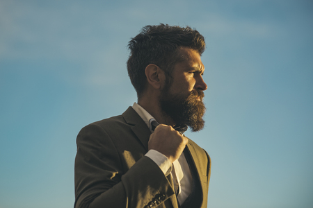 Guy with thoughtful face in luxury classic suit touches bow tie. Stylish hipster concept. Man with beard and mustache with dramatic sky on background. Hipster with stylish appearance in front of sky.の写真素材