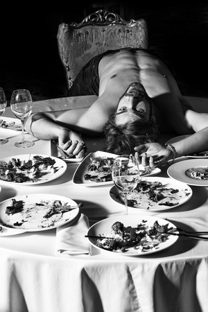 Handsome man or sexy muscular macho athlete bodybuilder with muscle torso undressed to waist lying on table with leftovers or residues food on dirty plates after banquet in restaurantの写真素材