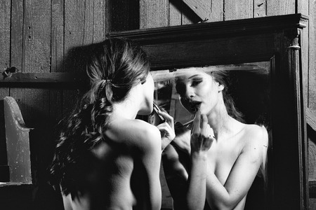 Sensual young lady with bare chest and curly hair near old dirty vintage mirror with lipstickの写真素材