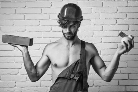 young handsome bearded macho man builder with sexy muscular athletic strong body has strong hands in orange hard hat or helmet holds pig dollar moneybox and brick on white wall backgroundの写真素材