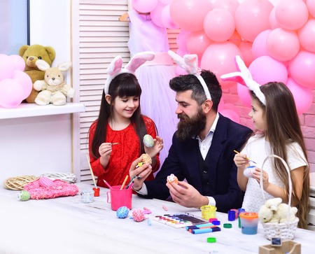 Father and daughters paint handmade egg. Easter, father and children in pink bunny ears. Happy...