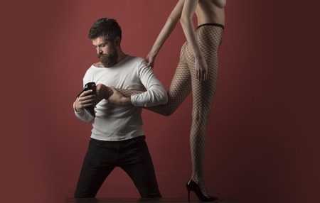 Man with strict face holds female leg and puts high heeled shoe on her foot. Lady in fishnet tights on burgundy background. Sexy Cinderella concept. Hipster found sexy naked Cinderella, copy space.の写真素材