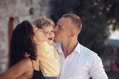 Happy family spend time together, urban background. Parents kissing son in cheeks, happy boy with smiling face. Mother and father love their child. Happy family concept. Parenthood, hugs, love, kiss.の写真素材