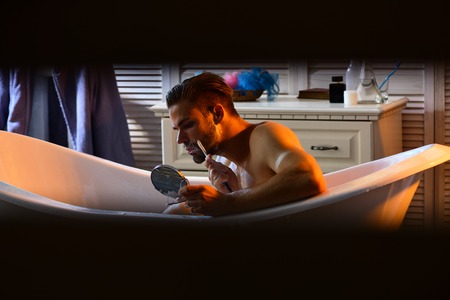 Macho with shaving soap on beard hair look in mirror. Bearded man shave with razor in bath in...
