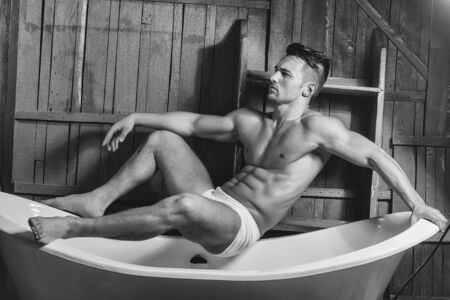 Handsome young man in white panties with sexy bare muscular torso sitting on bathtubの写真素材
