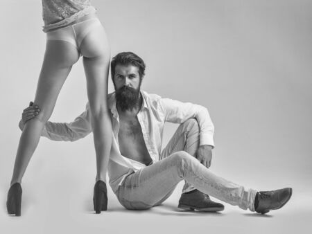 handsome bearded man with long beard on serious face sitting near sexy female legs and buttocks in lingerie and shoes of young pretty girl in studio isolated on white background, copy spaceの写真素材