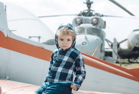 Cute boy sits on wing of old plane in aviation museum. Textured grunge old helicopter and sky with clouds on background. Kid on excursion to museum of aviation in open air. Air forces history concept.の写真素材