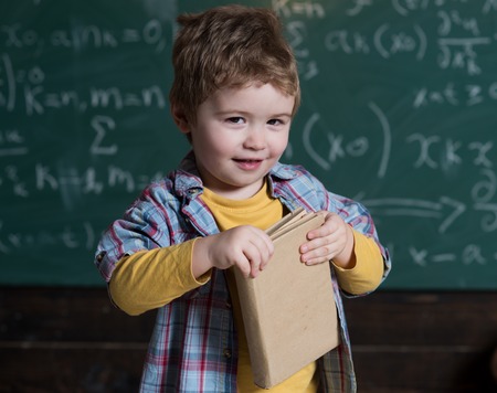 Smart child on smiling face, holds book in hands. Kid, preschooler or first former, chalkboard on background, defocused. Studying concept. Boy looks cute with book, kid likes to study.の写真素材